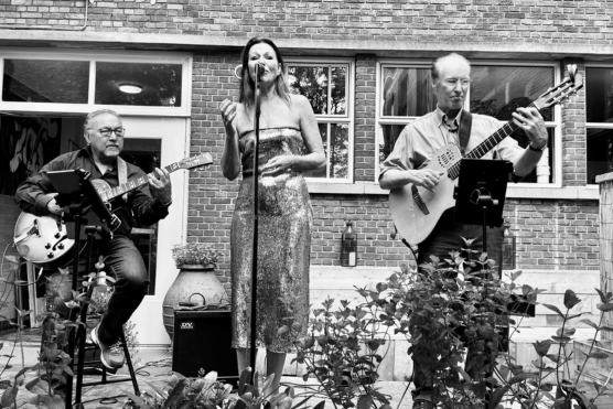 Links een man zittend op een kruk met een gitaar. In het midden een vrouw in een glitterjurk achter een microfoon. Rechts een staande man met een gitaar.